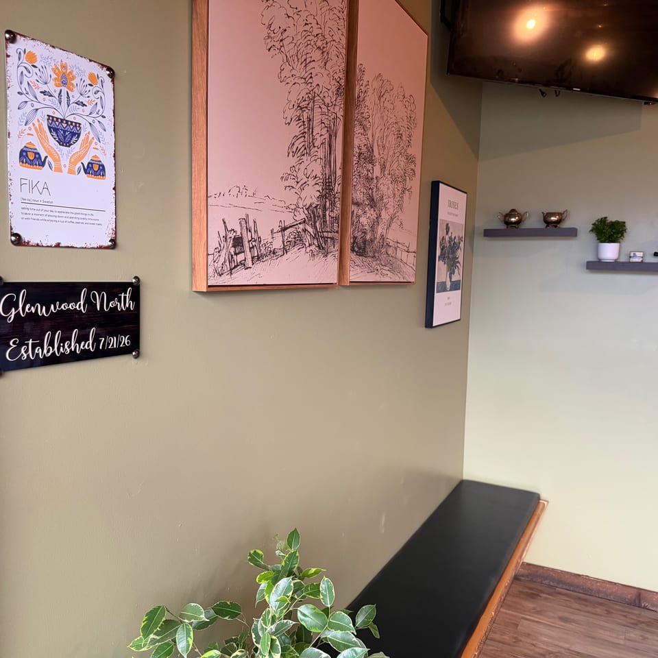 Restaurant interior featuring Fika signage, botanical artwork, decorative shelves, potted plants, and a wall-mounted television.