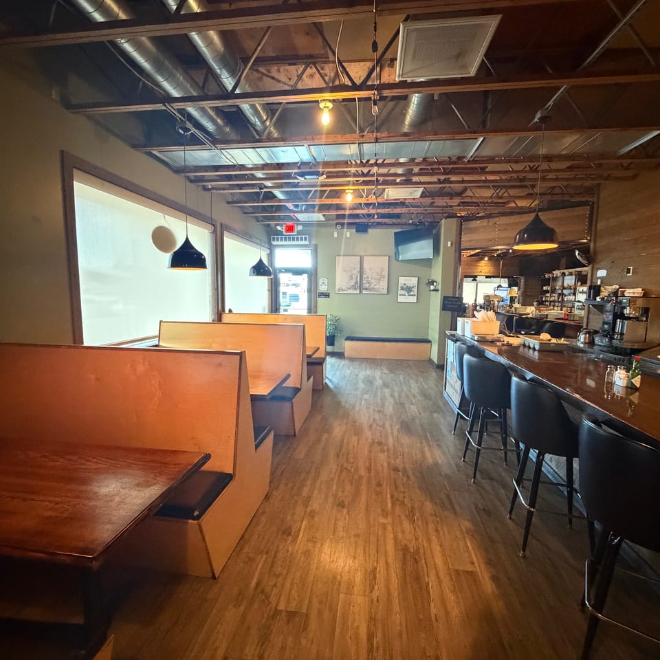 Warmly lit restaurant interior with wooden booths, bar seating, pendant lights, exposed ceiling ducts, and a service counter.