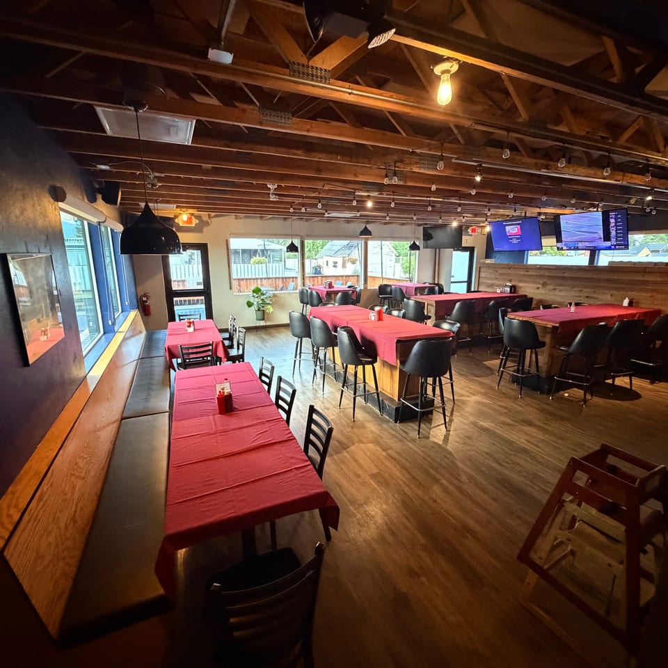Warmly lit restaurant dining room with red tablecloths, black chairs, exposed wooden beams, pendant lights, windows, and wall-mounted televisions.